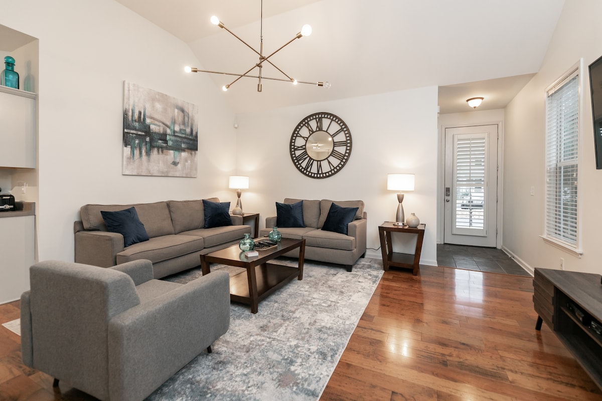 The living room features a spacious layout with comfortable seating arrangements and a modern coffee table. A large abstract art piece adorns the wall, complemented by a stylish wall clock. Natural light enters through a nearby door, illuminating the hardwood floors.