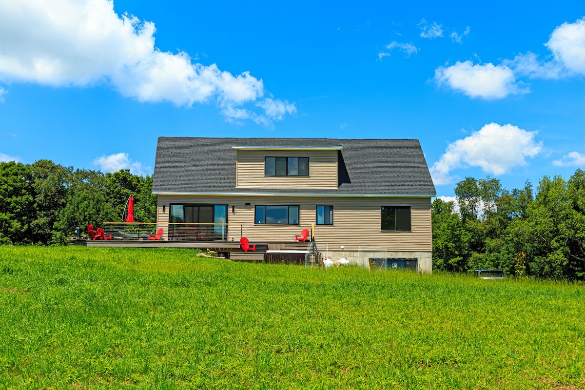 The modern cabin is set against a clear blue sky, surrounded by lush green grass. A spacious deck with bright red chairs extends from the house, offering outdoor seating. The building features multiple windows, emphasizing the connection with the natural landscape.