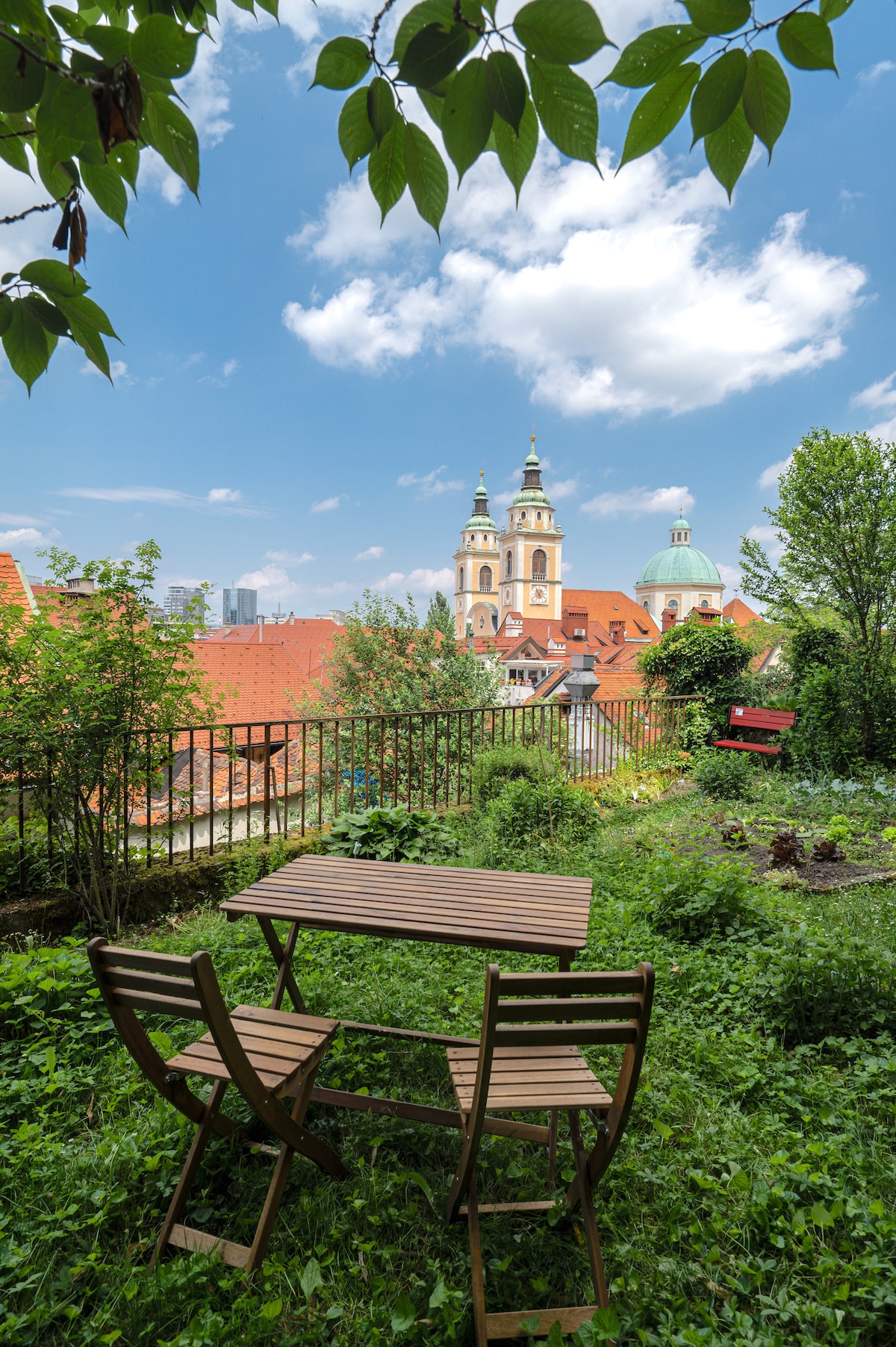 A secret garden oasis is displayed, featuring two wooden chairs arranged around a small table. Lush greenery surrounds the seating area, providing a sense of tranquility. The view includes rooftops and notable buildings, with church towers visible in the distance against a clear blue sky.