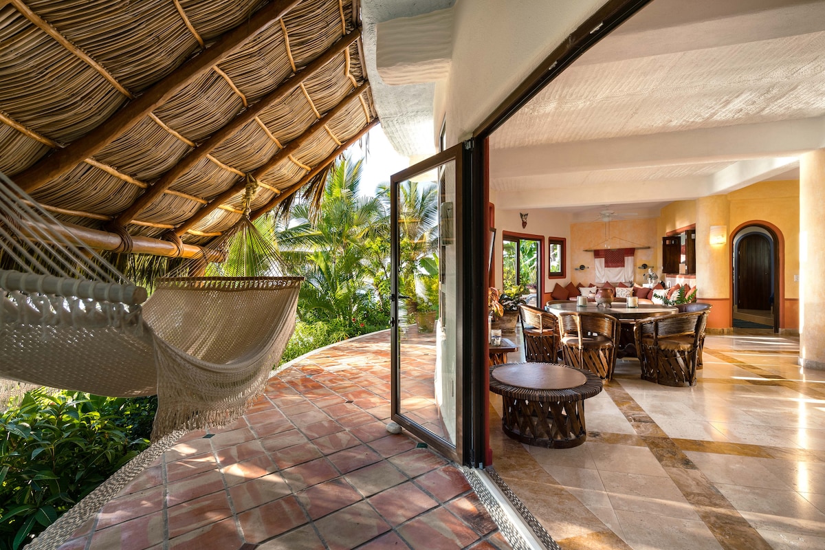 An outdoor terrace features a hammock suspended under a thatched roof, surrounded by lush greenery. A spacious living area is visible through large glass doors, showcasing a wooden dining table set for meals and inviting seating arrangements, offering a blend of comfort and natural elements.