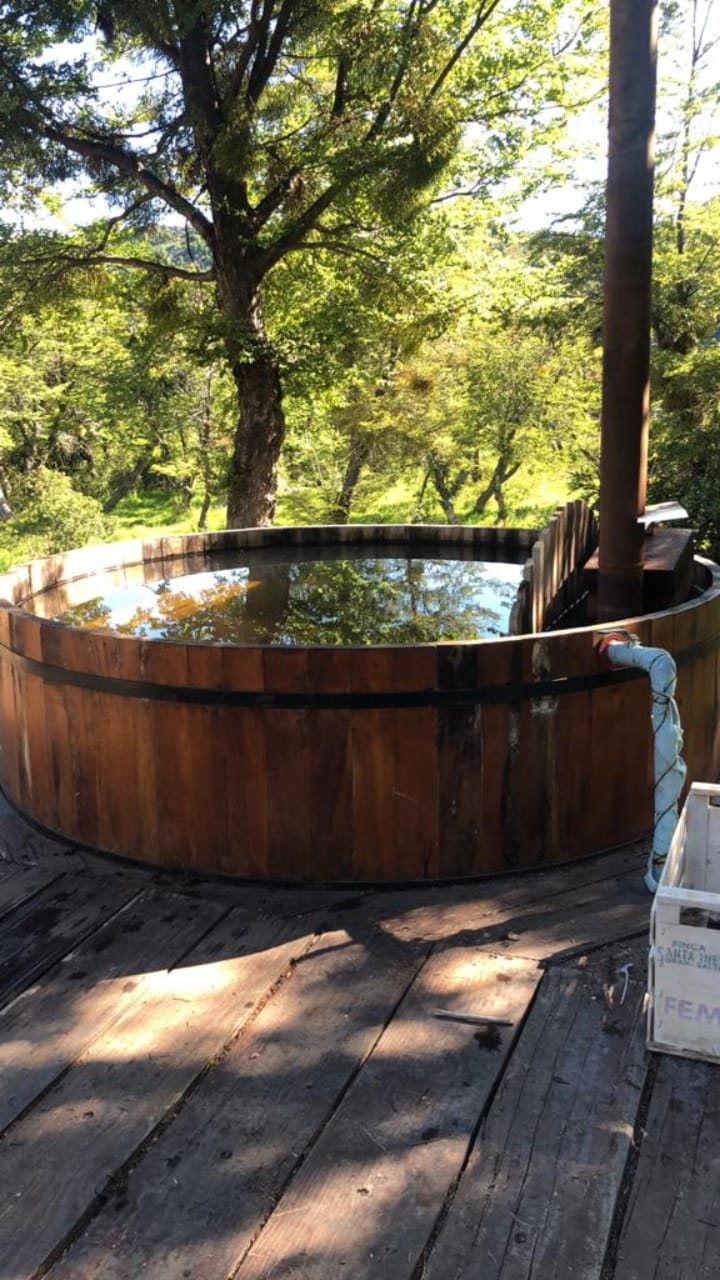 An exterior wooden hot tub is surrounded by lush greenery, providing a serene outdoor setting. The tranquil water reflects the trees above, while a chimney rises from one side, suggesting a cozy atmosphere for relaxation.