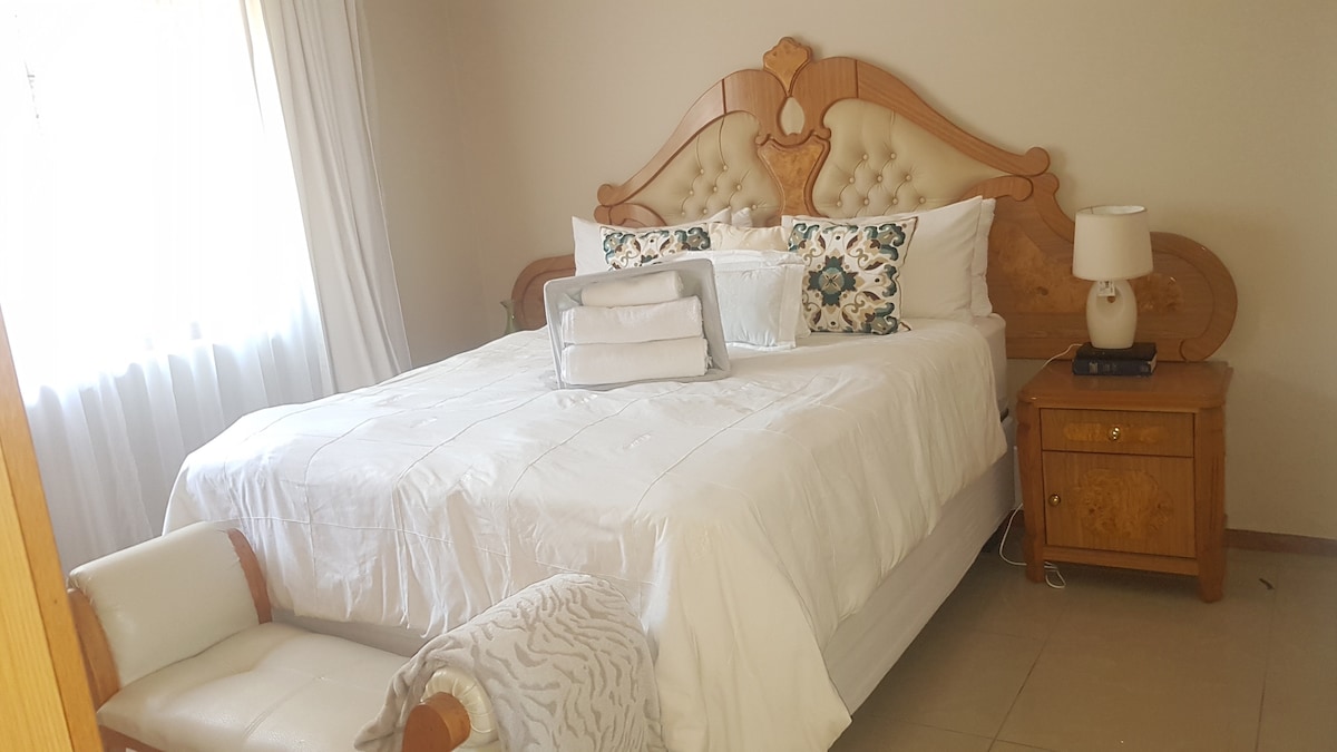 A spacious bedroom is featured with a well-appointed bed adorned in crisp white linens. Fluffy pillows and a decorative throw enhance the inviting appearance. A bedside table with a lamp provides soft lighting, and a comfortable chair is placed at the foot of the bed. Natural light filters through the window.