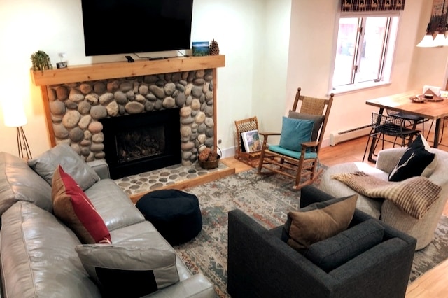 The living area showcases a remote-controlled gas fireplace framed by natural stone, enhancing the cozy atmosphere. Comfortable seating includes a gray sofa and armchair, with colorful accent cushions. A wooden rocking chair and dining table are visible, creating a relaxing gathering space.