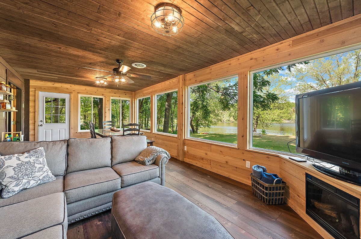 A sunlit room features large windows that offer views of the surrounding landscape, including the river. A sectional couch and an ottoman provide comfortable seating, while a television is positioned nearby. The space is adorned with wooden walls and a ceiling that adds warmth to the atmosphere.