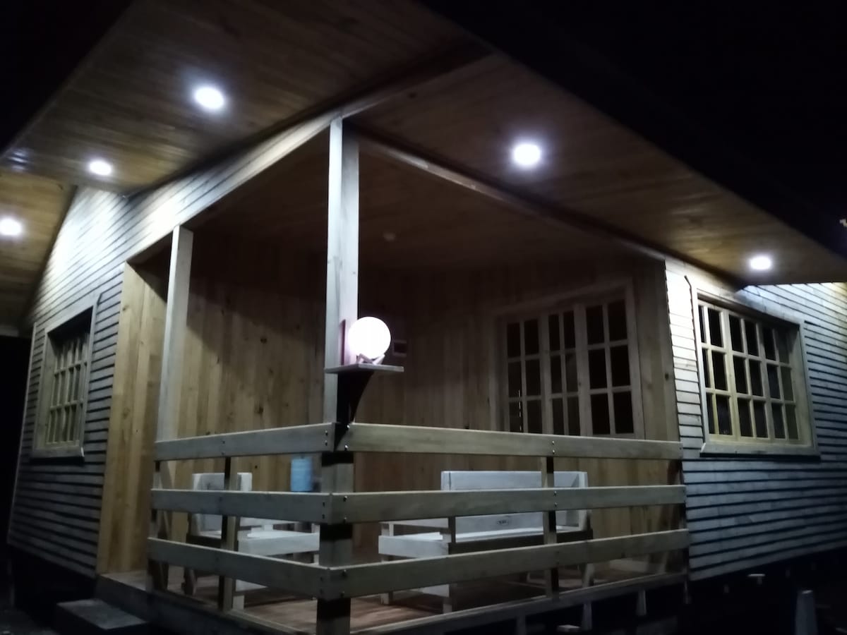 The exterior of a wooden cabin is illuminated at night, showcasing its welcoming porch. Soft lighting highlights the wooden texture of the siding and beams, while multiple windows with paned glass offer a glimpse into the warm interior.
