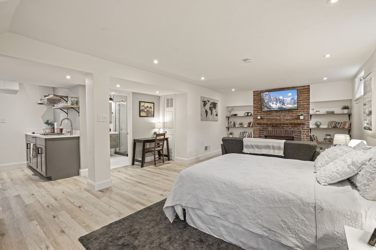 A spacious basement studio features a queen bed with soft white linens, complementing a light, neutral color scheme. A cozy seating area includes a couch near a modern wall-mounted TV. The small kitchen is visible, highlighted by sleek cabinetry and a dining table for two.