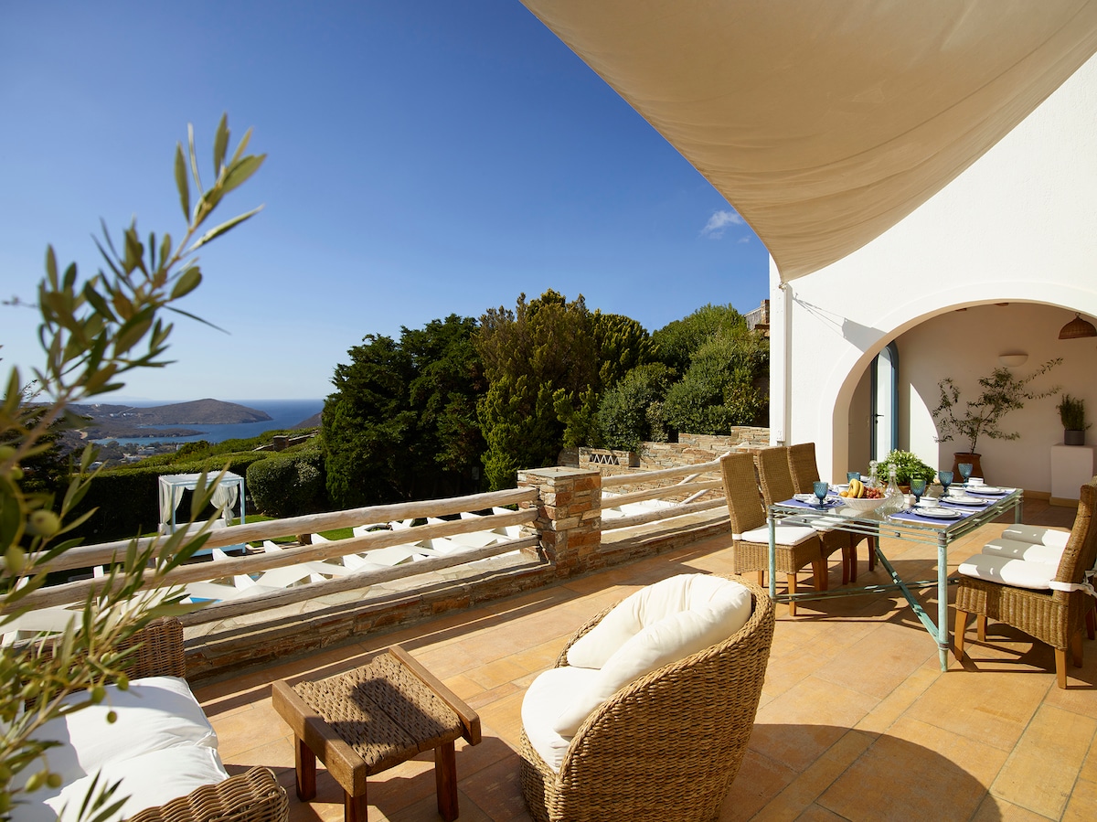 An outdoor terrace is highlighted by comfortable seating arrangements, complemented by a table set for dining. A distant view reveals the blue hues of the sea and lush greenery, while natural light brightens the space under a beige awning.