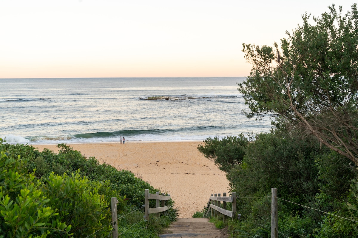 A sandy path leads down to a serene beach, flanked by lush greenery on both sides. Gentle waves roll onto the shore, and a pair of figures can be seen walking along the water's edge, set against a soft pastel sky.