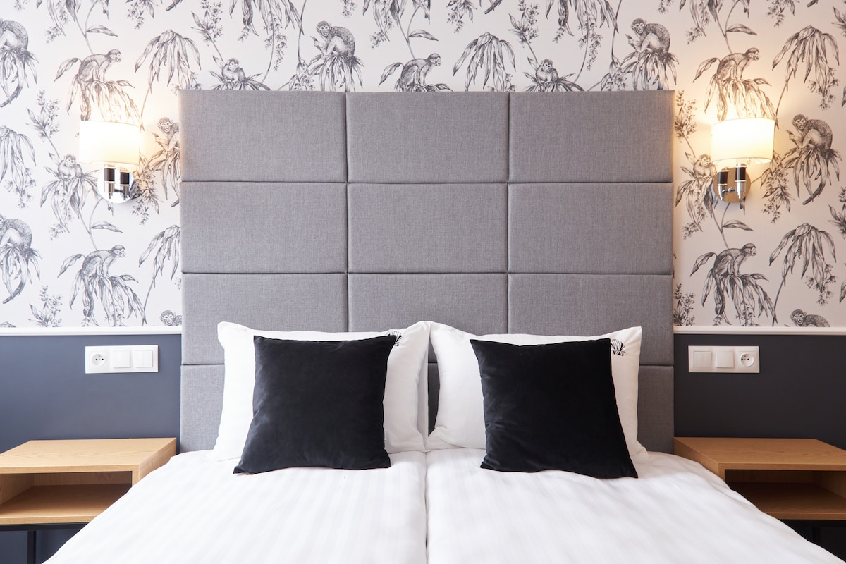 A modern room features a spacious bed with a gray tufted headboard. Two black decorative pillows are positioned on the white bedding. Soft lighting from wall sconces complements the floral wallpaper, providing a soothing ambiance.