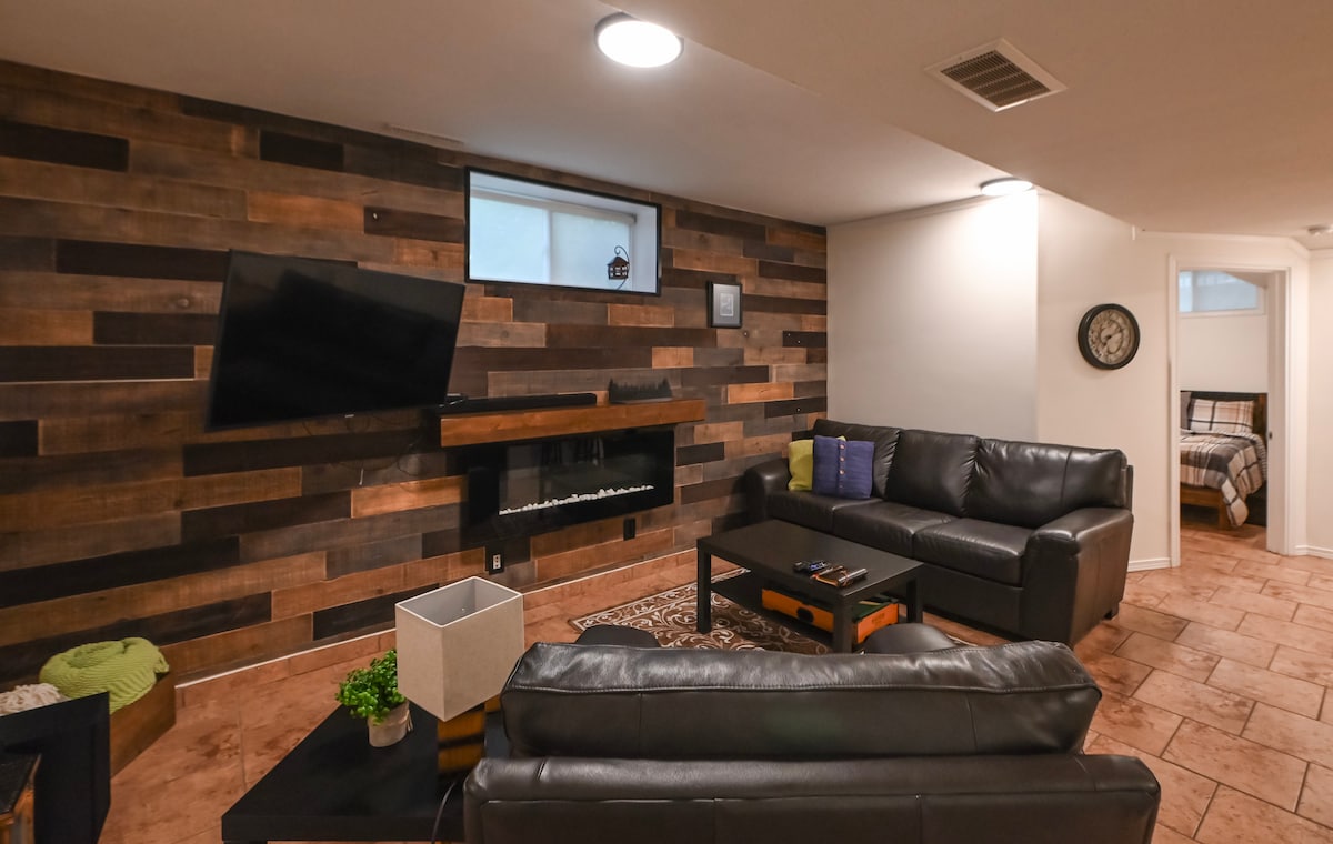 The living area showcases a comfortable leather sofa facing a wall-mounted television. A modern electric fireplace is positioned beneath it, complemented by rich wooden paneling. A small table and decorative plant are visible, contributing to the inviting ambiance.