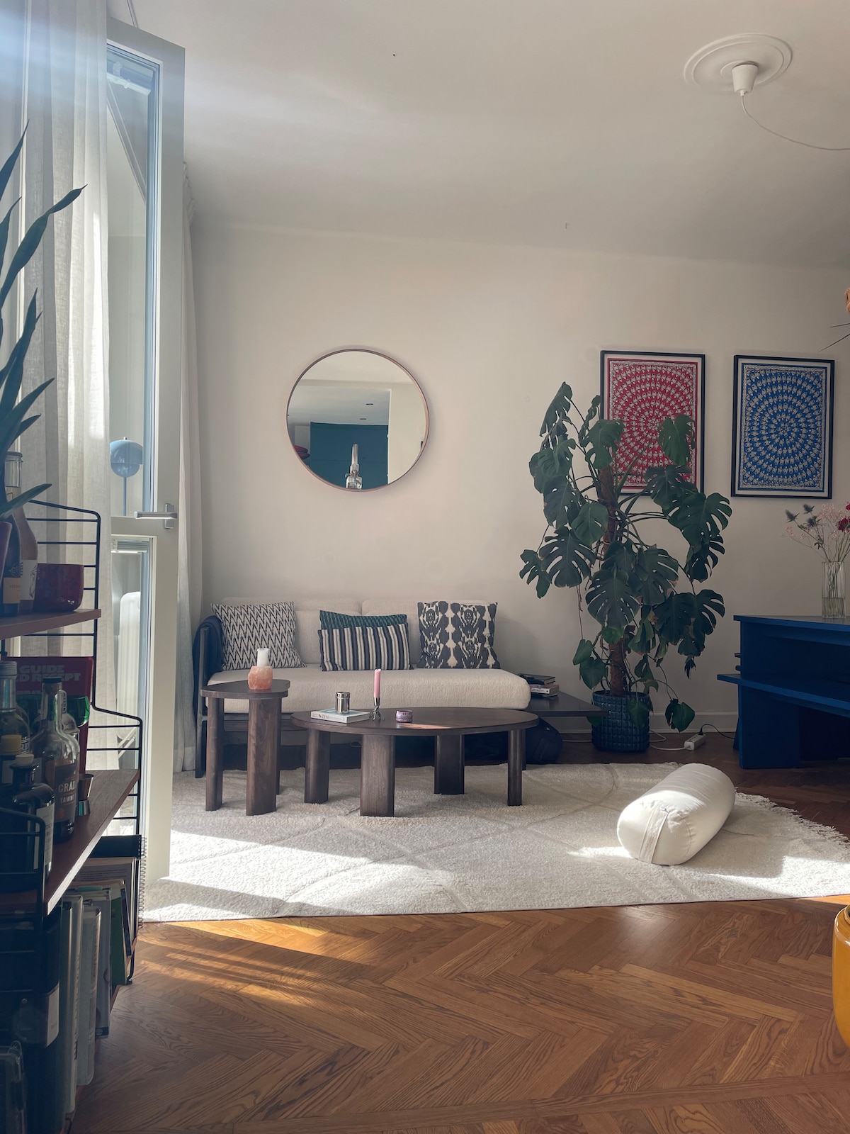 A cozy living room features a light-colored three-seat sofa with patterned cushions, complemented by a round coffee table. A large indoor plant adds greenery, while sunlight streams in through big windows, creating a warm atmosphere. Art pieces adorn the wall, enhancing the space.