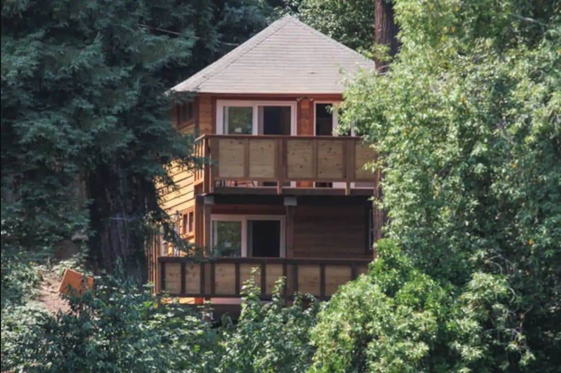 The exterior of a charming wooden duplex is showcased, nestled among lush green trees. Multiple balconies are visible, providing access to outdoor seating and views of the surrounding nature. The upper and lower levels are designed with ample windows to invite natural light.
