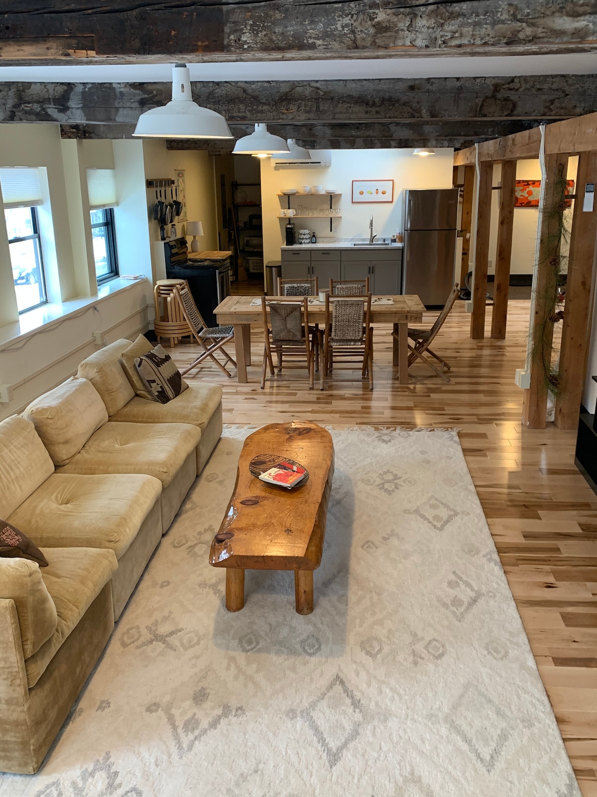 A spacious living area features a comfortable sofa and a wooden coffee table. Natural light enters through large windows, illuminating the hardwood flooring. In the background, a dining area with a table and chairs is visible, along with a kitchen equipped with modern appliances.