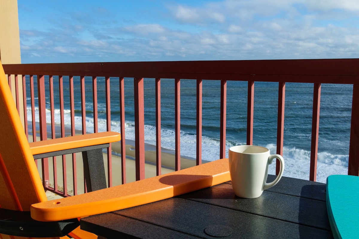 A private balcony features bright orange adirondack chairs facing the ocean. A white mug rests on a dark outdoor table, with the waves gently rolling onto the shore in the background under a partly cloudy sky.