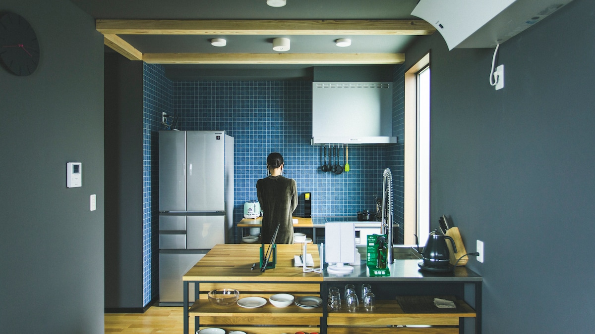 A modern kitchen features a wooden dining table in the foreground, surrounded by plates and glassware. A person stands at the countertop, with a large refrigerator and stove visible in the background against blue tiled walls, offering ample workspace and natural light from the window.