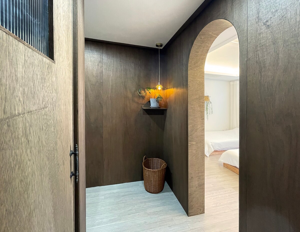 A hallway features warm wood paneling and a minimalist design. A decorative shelf holds a small plant and a sculpture, while a woven basket sits on the floor. An archway leads to a bright sleeping area, enhancing the inviting walk-through space.