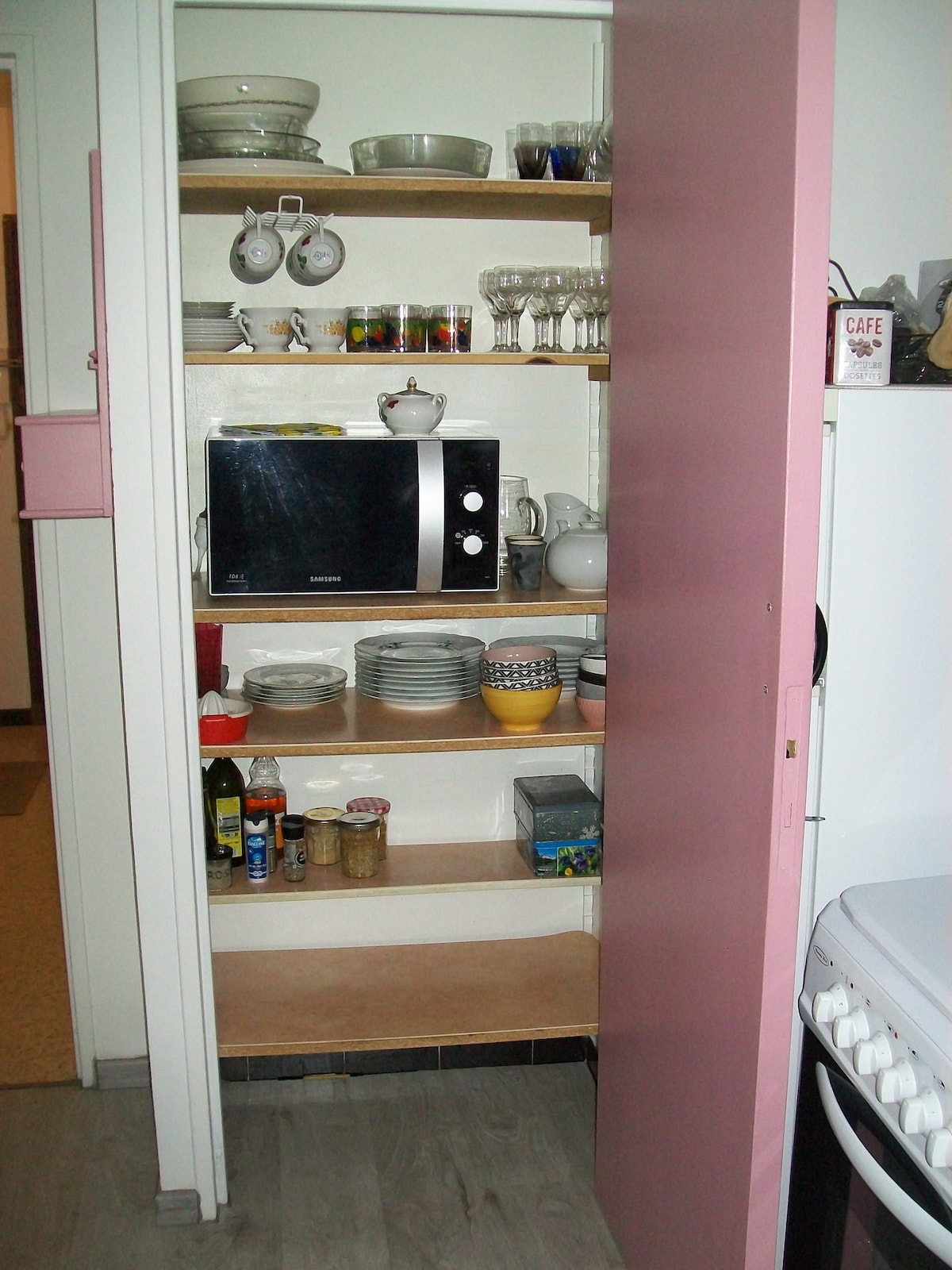 A compact kitchen cabinet contains various items, including plates, glasses, and a microwave. Shelves are organized, showcasing a variety of cookware and utensils. Light-colored wood enhances the visibility of the items, while a pink door adds a subtle touch of color.