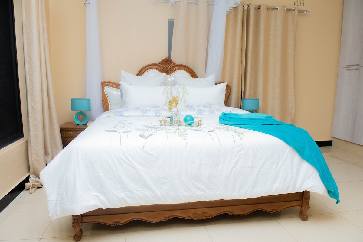 A spacious bed is positioned in the center of the room, featuring a white linens adorned with floral designs. Coordinating bedside lamps are placed on each side. A soft turquoise throw is elegantly draped across the bed, complementing the light beige walls and window drapes.