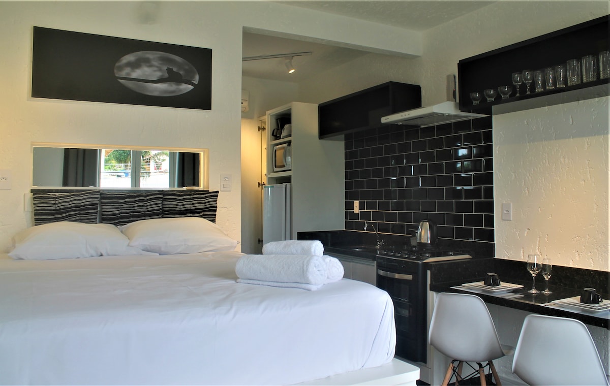 A cozy studio space features a spacious bed with crisp white linens and a modern kitchen area. Dark tiles complement the sleek countertop, while utensils are neatly arranged on open shelving. Natural light illuminates the room, creating a calming atmosphere.