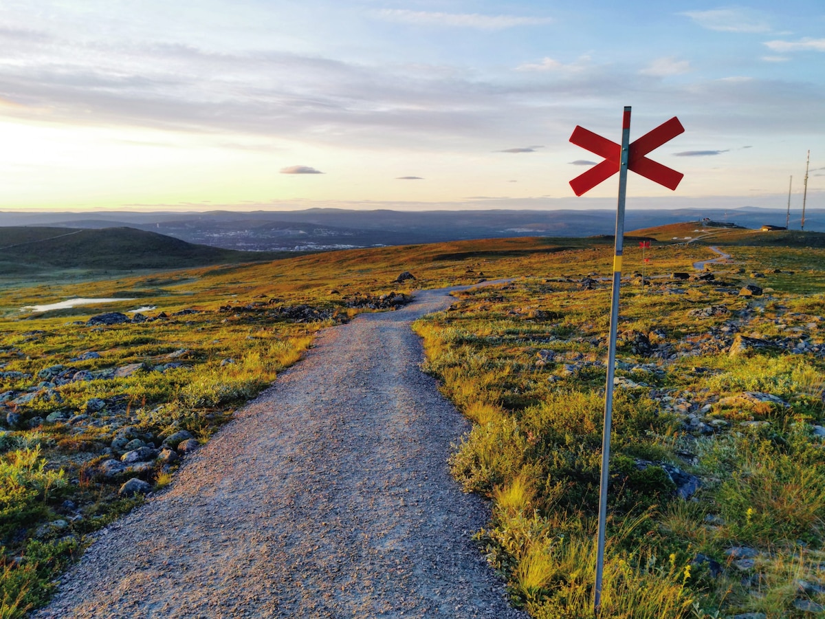 A gravel path meanders through a grassy landscape, marked by a distinctive red X sign. Rolling hills and distant mountains create a serene backdrop, with a clear sky that transitions from soft yellow to blue, suggesting a peaceful outdoor environment for exploration.