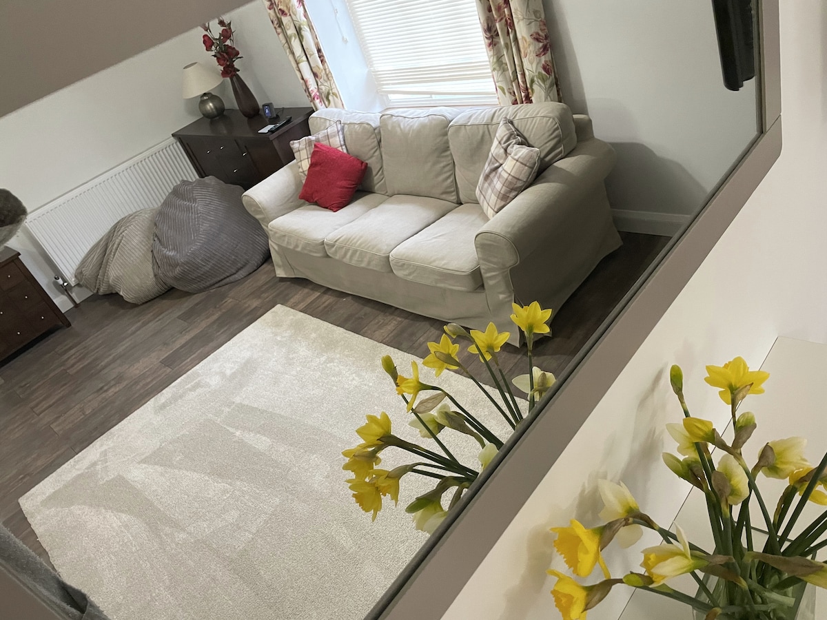 A spacious sitting room is viewed through a reflective surface, showcasing a cream-colored sofa with a red accent pillow. A light area rug is placed on the floor, while a vase of yellow flowers sits in the foreground. Natural light illuminates the room.