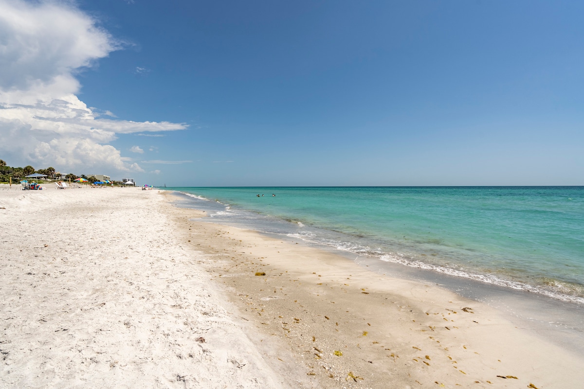 A serene beach scene showcases soft white sands meeting clear turquoise waters. Gentle waves lap at the shore under a bright blue sky, with a few scattered clouds. Sunbathers can be seen in the distance, enjoying the tranquil environment.