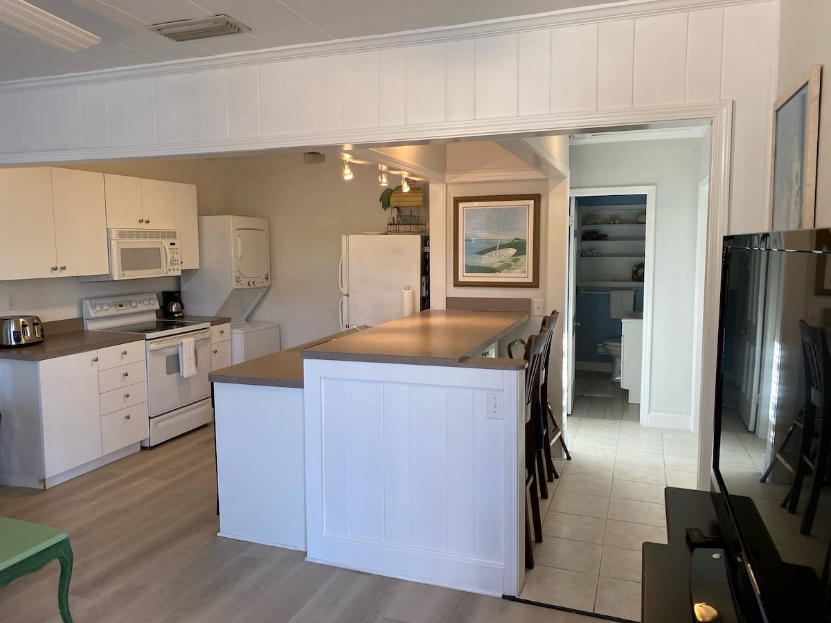 An open kitchen area features a large island with seating and ample counter space. Light-colored cabinets provide storage, while modern appliances, including a refrigerator and washer-dryer, are visible. The living room is adjacent, with a flat-screen TV positioned against a white wall.
