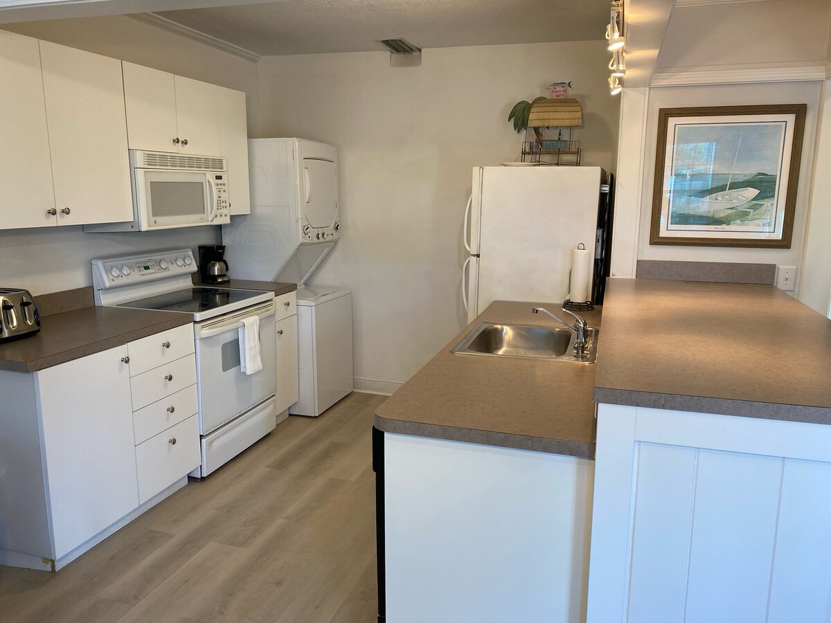 The kitchen is equipped with modern appliances, including a microwave, washer, dryer, and refrigerator. Counter space surrounds a stainless-steel sink, with ample cabinets for storage. A subdued color palette and natural light enhance the open layout, creating a functional cooking area.