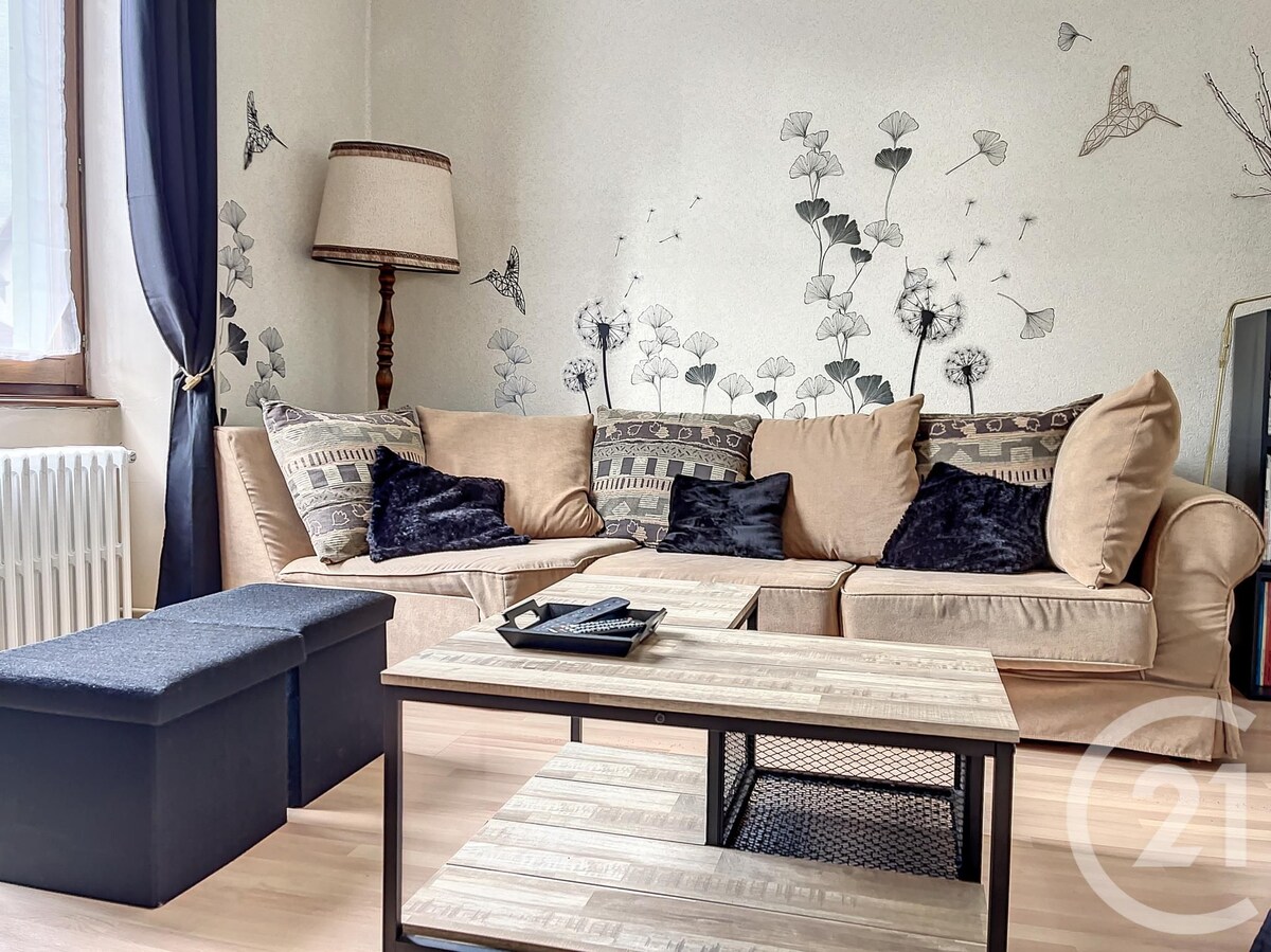 A comfortable living area is presented with a beige sofa adorned with dark cushions. A coffee table made of wood and metal sits in front, while a floor lamp provides illumination. The walls feature botanical designs that add a subtle elegance to the space.