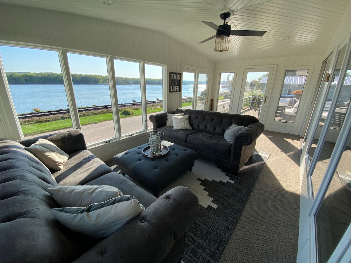 A sunlit three-season room features large windows providing views of the Mississippi River. Two plush sofas are arranged comfortably on a defined area rug, complemented by decorative pillows. A ceiling fan maintains airflow, and a decorative lantern adds a touch of charm to the space.