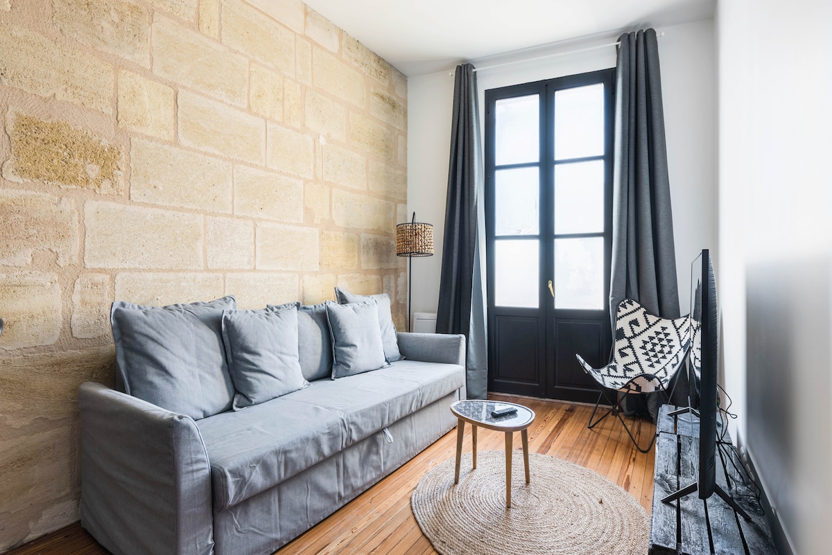 A cozy living area is highlighted by a comfortable gray sofa accompanied by a round wooden coffee table and a patterned accent chair. Natural light filters through large glass doors, framed by flowing gray curtains. A flat-screen television is positioned beside a textured stone wall.