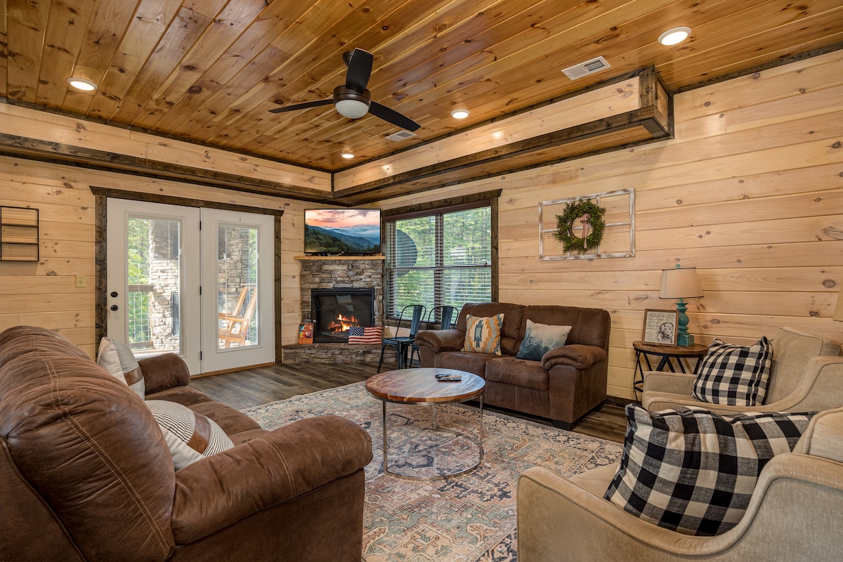 The living room features wood-paneled walls and a ceiling with recessed lighting. A comfortable seating arrangement includes a brown leather couch and an armchair. A round coffee table sits on a patterned rug, with an electric fireplace and a TV mounted on the wall.