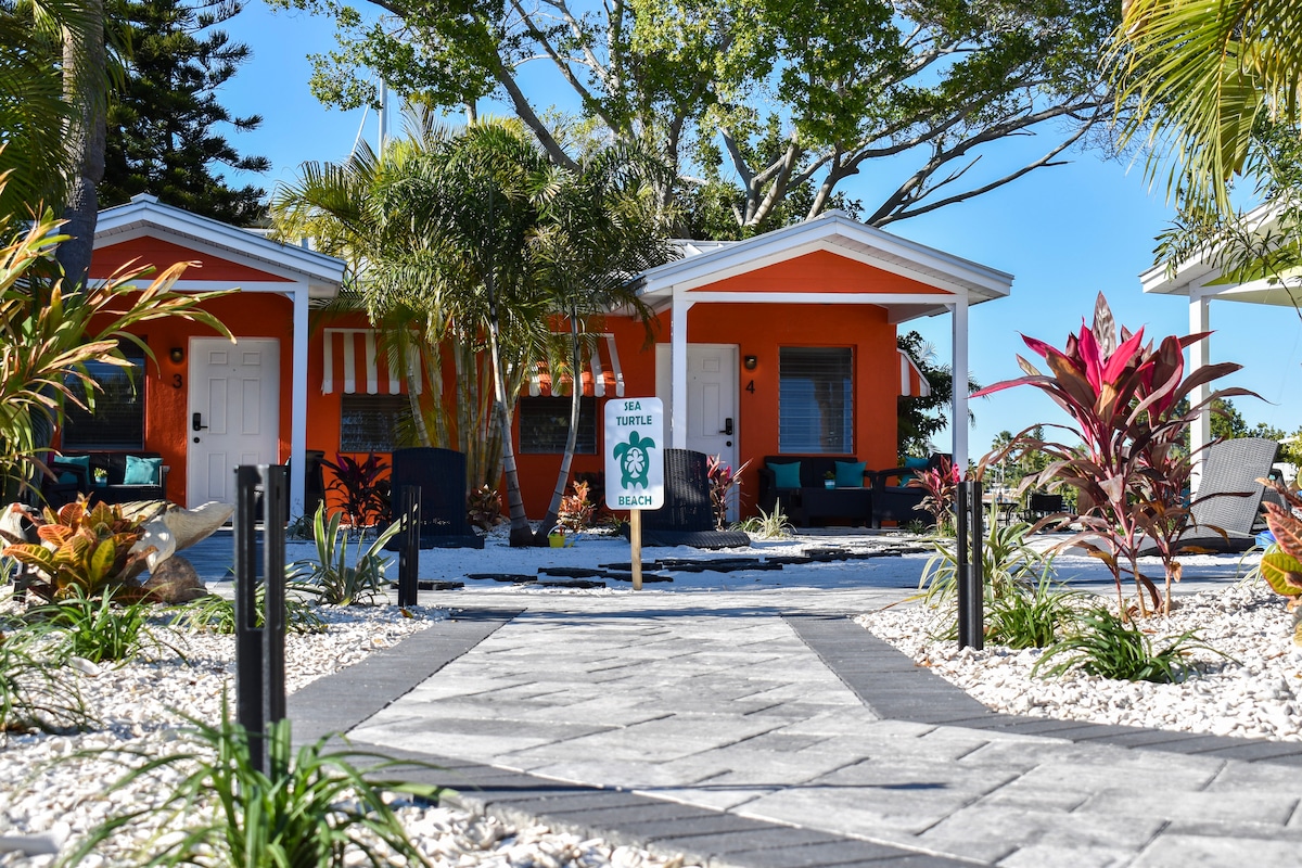 A vibrant orange bungalow is framed by tropical landscaping, including lush green plants and colorful flowers. A stone pathway leads to the entrance, guiding guests towards a welcoming atmosphere. The clear blue sky complements the lively exterior, enhancing the inviting appearance of the property.