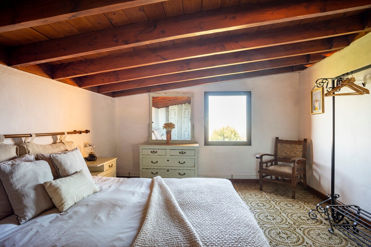 A spacious bedroom is captured, featuring a large bed adorned with soft pillows. Natural light filters through a window, illuminating a vintage mirror and a wooden dresser. An antique chair complements the inviting decor, while exposed wooden beams enhance the room's rustic charm.
