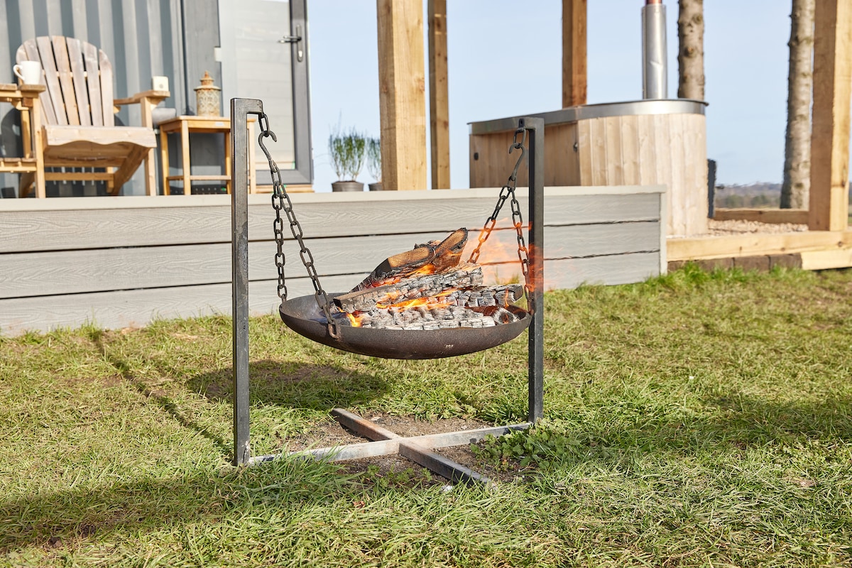 A suspended fire pit is seen in the foreground, with flames dancing above glowing embers. Nearby, wooden chairs are positioned, complementing a cozy outdoor space. In the background, a wood-fired hot tub is visible, surrounded by grassy areas and structures that enhance the tranquil setting.