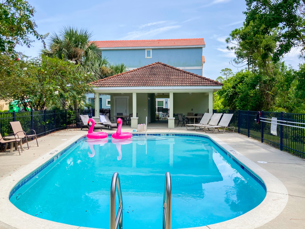 A clear, inviting swimming pool is framed by a tiled deck and surrounded by lush greenery. Several lounge chairs are positioned nearby for relaxation. Two pink inflatable flamingos float gently on the water's surface, while a covered patio area is visible in the background.