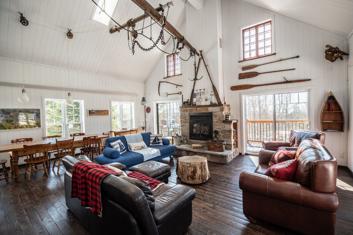 A spacious open-concept living area is presented, featuring cozy seating arrangements with a blue sofa and brown leather couches. A stone fireplace serves as a focal point, while large windows allow natural light to illuminate the space. Rustic decor, including wooden paddles and wall hooks, enhances the cabin's charm.