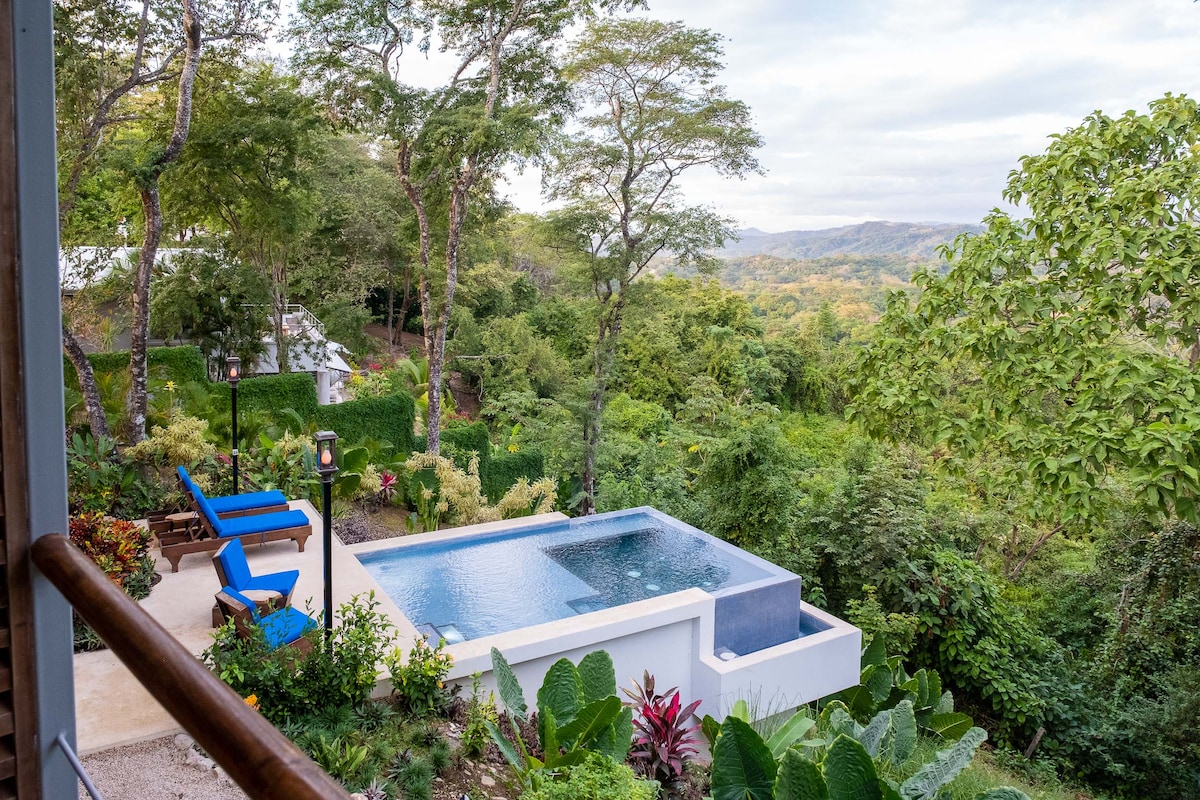 An infinity pool is nestled within a lush garden, surrounded by vibrant greenery. Comfortable lounge chairs are positioned nearby, offering a tranquil view of the surrounding hills and trees. The serene landscape showcases a blend of tropical plants and distant mountains.