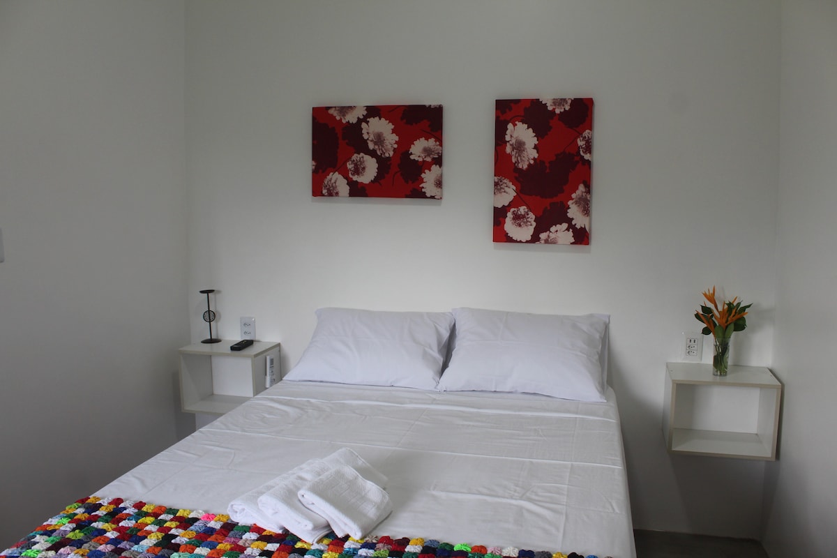 A clean and bright sleeping area features a spacious bed with crisp white linens. Colorful wall art hangs above the bed, complemented by two bedside tables with modern lamps. Fresh towels are neatly arranged on the bed, providing a welcoming touch.