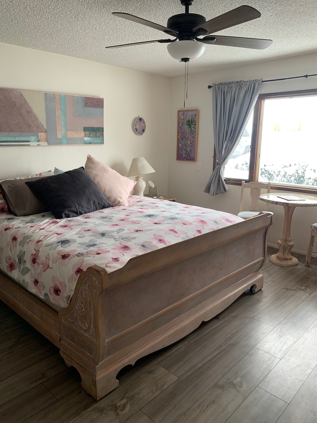A private bedroom features a queen bed with floral bedding and several accent pillows. A window allows natural light to fill the space, and a small table with two chairs is positioned nearby. Soft curtains frame the window, adding to the calming atmosphere.
