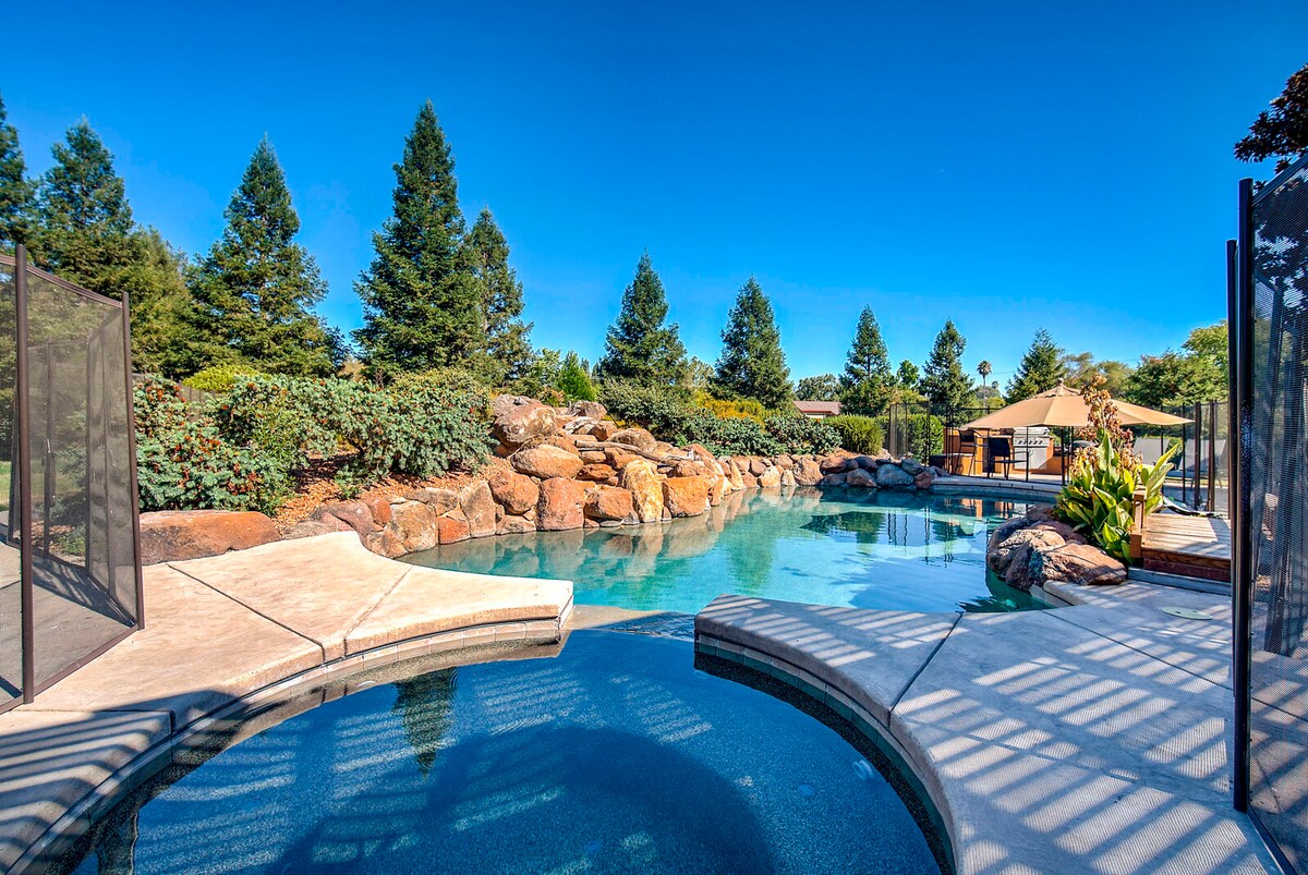 A serene outdoor area features a large, unheated swimming pool surrounded by natural stone. The pool is bordered by lush greenery, adding to the tranquil ambiance. A safety fence encloses the hot tub, providing an inviting space for relaxation.