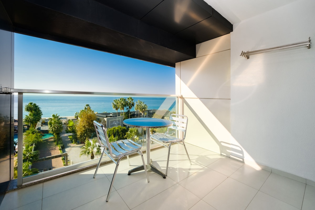 A balcony area features a round table with two chairs, providing a view of the blue ocean and surrounding palm trees. Natural light brightens the space, which is enhanced by a sleek glass railing. A clothing rack is installed for convenience.