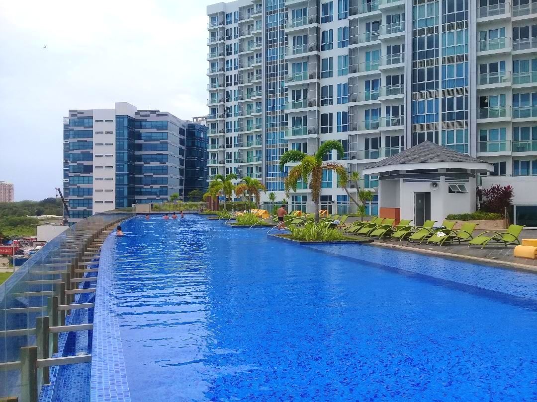 An expansive infinity pool is seen alongside a modern building. Lounge chairs are arranged along the pool's edge, and palm trees add greenery to the surroundings. The clear blue water reflects the sky, creating a tranquil atmosphere for relaxation.