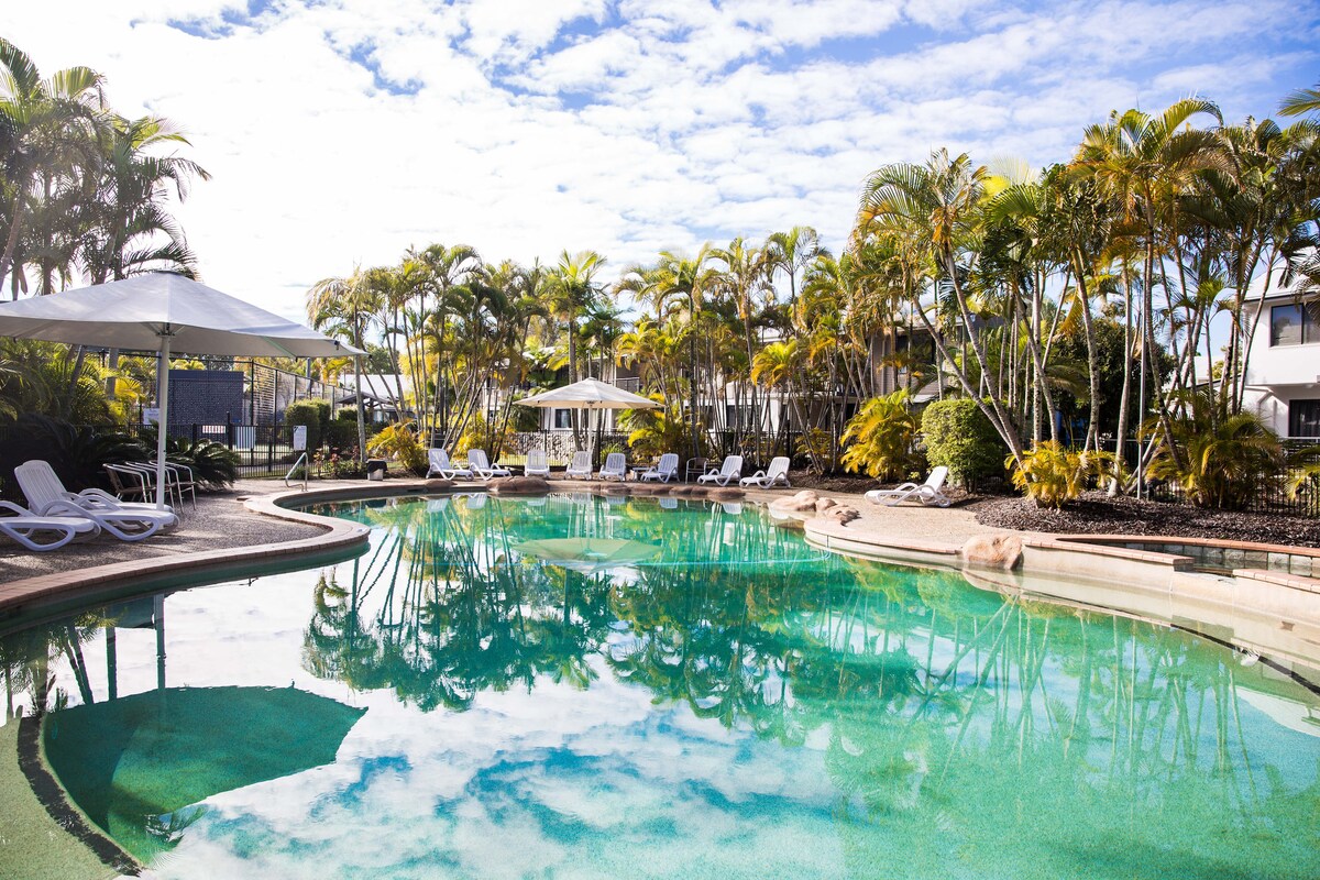 A serene swimming pool is surrounded by palm trees, reflecting the sky. Sun loungers are placed around the pool, providing spaces for relaxation. Umbrellas offer shade for guests enjoying the tranquil environment filled with lush greenery.