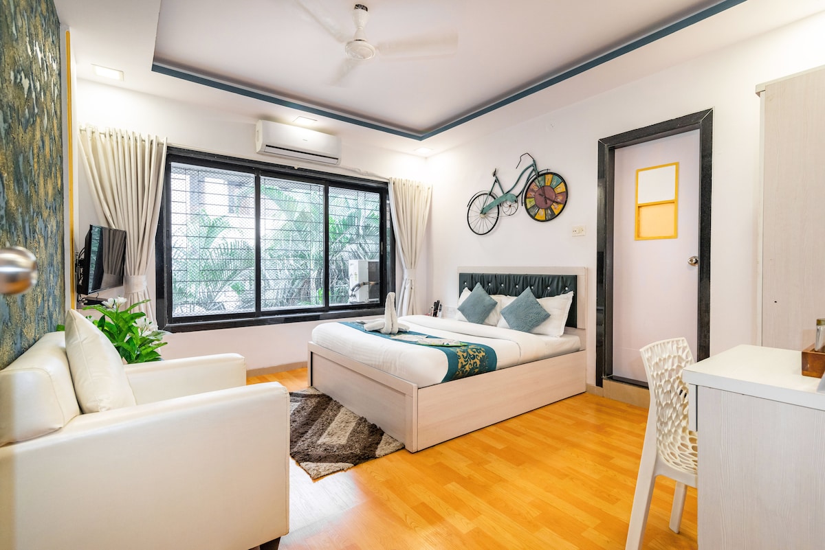 A spacious bedroom is furnished with a comfortable bed and a cozy sitting area. Large windows allow natural light, offering views of greenery. A decorative bicycle is mounted on the wall, and a small dining table is present, enhancing functionality.
