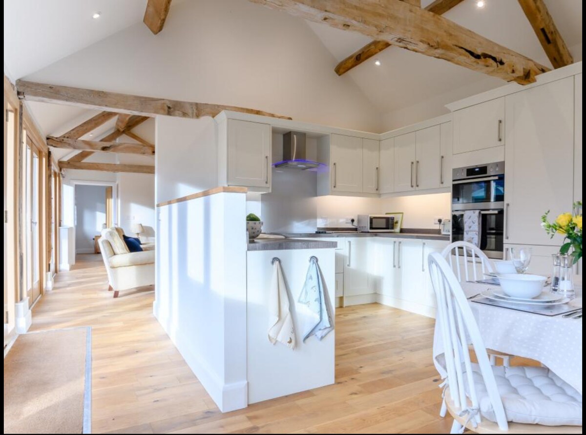 A spacious kitchen area showcases modern appliances, including an oven and microwave, set against light cabinetry. Wooden beams accentuate the high ceiling. A dining table is arranged with tableware and flowers, while a comfortable seating area is visible in the background through large windows.