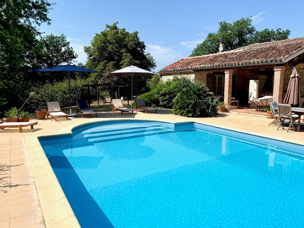 A clear view of a salt-water pool is presented, surrounded by sun loungers and parasols. The property features a stone exterior with a tiled roof, complemented by greenery and trees in the background. A spacious terrace area invites relaxation and social gatherings.