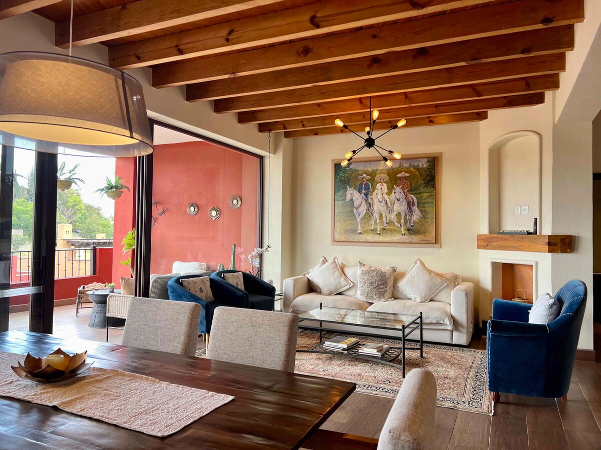 The open-concept living and dining area features wooden beams overhead, with large windows allowing natural light. Comfortable seating is provided by a mix of plush sofas and chairs, accompanied by a decorative area rug and a dining table that enhances the inviting space.