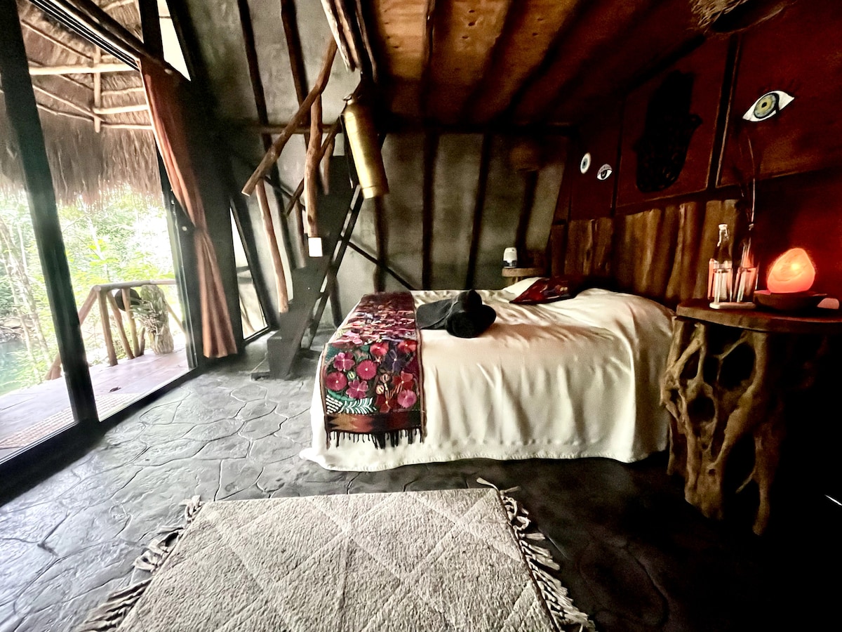 A cozy room features a spacious bed dressed in a soft white coverlet. Natural textures dominate the decor, complemented by a unique wooden side table. Large glass doors provide a view of the surrounding greenery and access to a private outdoor space.