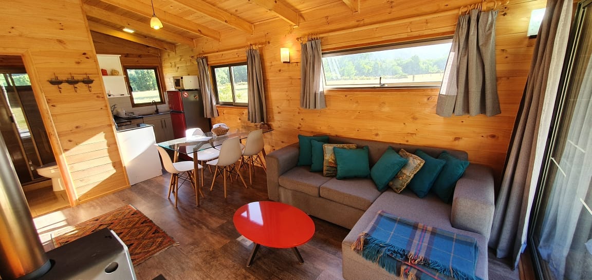 A cozy living area is highlighted by a modern grey sofa adorned with turquoise cushions. A vibrant red coffee table sits in front, accompanied by a light wood dining table with white chairs. Large windows allow natural light to flood the space, showcasing the surrounding landscape.